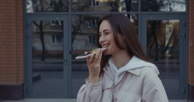 Gülümseyen genç kadın caddede duruyor ve modern akıllı telefondan ses kaydı yapıyor. Açık havada sesli mesaj bırakmak için cep telefonu kullanan çekici bir esmer..