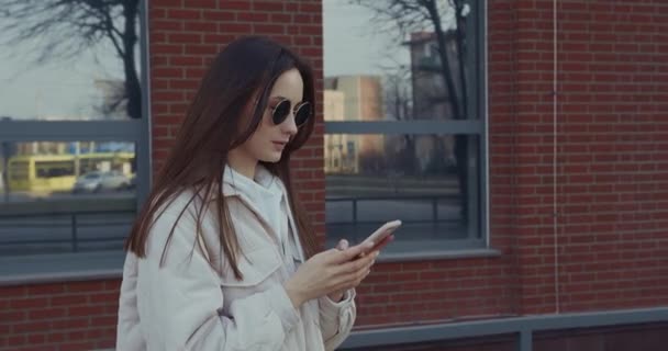 Femme agréable portant des vêtements décontractés et des lunettes de soleil rondes naviguant sur Internet sur smartphone moderne. Jolie femme marchant sur la rue de la ville avec gadget sur les mains. — Video