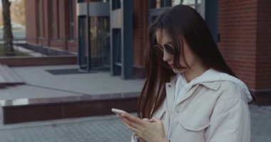 Gündelik giyinen, güneş gözlüklü, modern akıllı telefonu elinde tutan hoş bir genç kadın. Teknoloji ve yaşam tarzı kavramı.