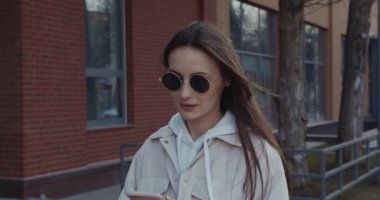 Elinde kişisel akıllı telefonuyla caddede yürüyen düz kahverengi saçlı olumlu bir kadın. Güneş gözlüklü çekici bayan modern aletleri açık havada kullanıyor..