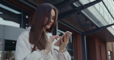 Kahverengi saçlı güzel bir bayan caddede duruyor ve kişisel akıllı telefon kullanıyor. Modern aletler, boş zaman ve yaşam tarzı kavramı.