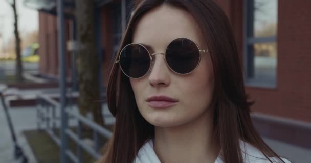 Приголомшлива темноволоса жінка в повсякденному одязі і круглих сонцезахисних окулярах, що стоять на міській вулиці. Концепція людей, молоді та способу життя . — стокове відео
