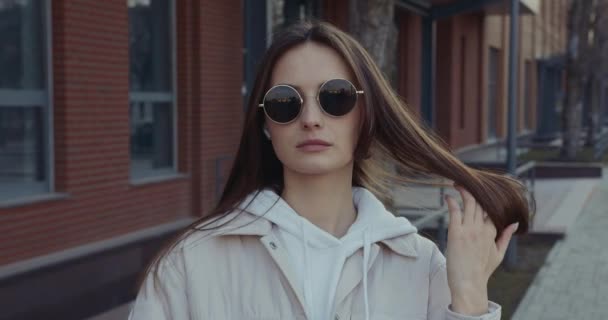 Ελκυστική γυναίκα με γυαλιά ηλίου και casual φόρεμα στέκεται στο δρόμο, αγγίζοντας καστανά μαλλιά και κοιτάζοντας την κάμερα. Νεαρή γυναίκα περνάει ελεύθερο χρόνο σε εξωτερικούς χώρους. — Αρχείο Βίντεο