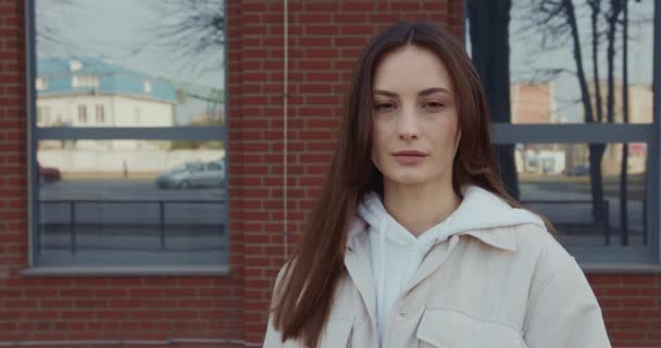 Retrato de uma jovem encantadora com cabelo castanho posando no espaço urbano. Adorável fêmea vestida com roupas casuais em pé ao ar livre e olhando para a câmera. — Vídeo de Stock