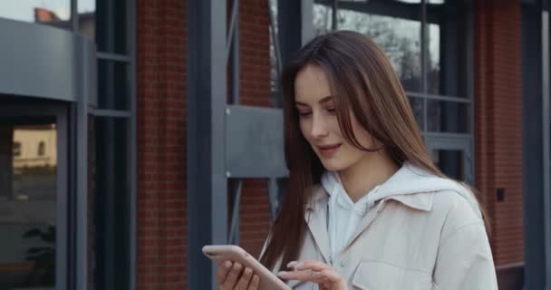 Attraktive junge Frau mit braunen Haaren surft auf dem Smartphone im Internet, während sie auf der Straße spaziert. Konzept von Menschen, Technologie und Lebensstil. — Stockvideo