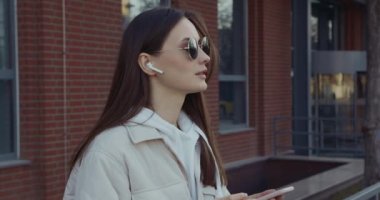 Dışarıyı aramak için kablosuz kulaklık ve akıllı telefon kullanan çekici bir kadın. Güneş gözlüklü genç esmer, gülümsüyor ve temiz havada konuşuyor.. 