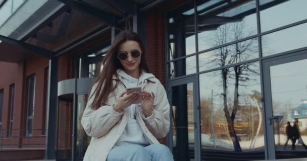 Positiv kvinna i solglasögon och casual outfit sitter nära modern byggnad och skriva meddelanden på modern smartphone. Begreppet avkoppling och livsstil. — Stockvideo