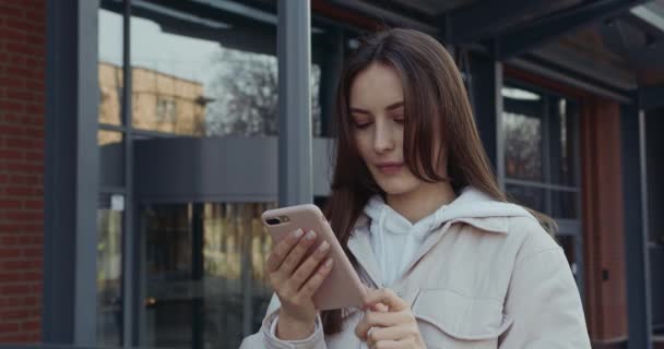 Portrét krásné šťastné ženy v ležérní oblečení stojící venku a pomocí moderního smartphonu. Hezká brunetka tráví volný čas na čerstvém vzduchu s osobní gadget v rukou. — Stock video
