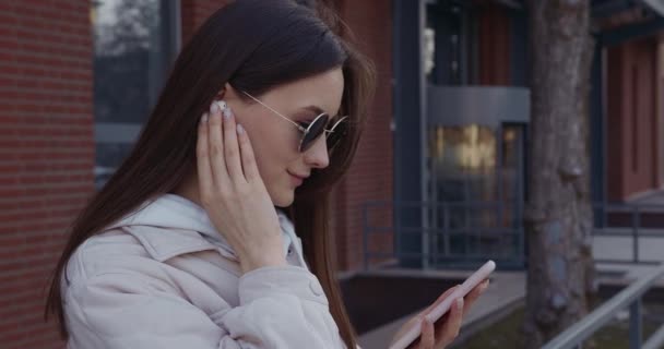 Imádnivaló nő okostelefonnal a kezében zenét hallgat vezeték nélküli fülhallgatóval. Fiatal barna napszemüveg áll az utcán, és a modern szerkentyű. — Stock videók
