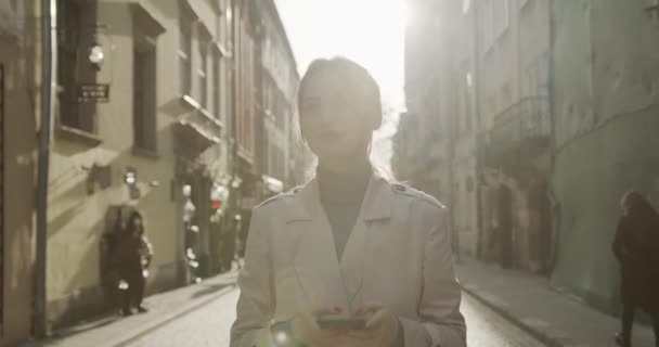 Attraktive junge Frau mit braunen Haaren surft auf dem Smartphone im Internet, während sie auf der Straße spaziert. Konzept von Menschen, Technologie und Lebensstil. — Stockvideo