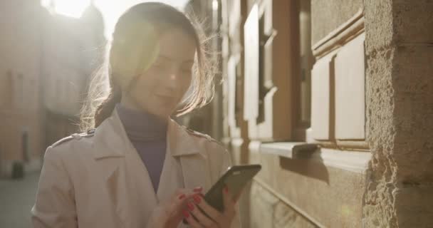 Leende ung kvinna med rakt mörkt hår står på stadens gata och sms: a meddelanden på smartphone. Bedårande dam i avslappnad outfit med mobil för online-kommunikation. — Stockvideo