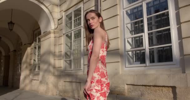 Modelo de moda joven con el pelo castaño liso posando cerca del edificio viejo en la calle de la ciudad. Encantadora mujer con vestido de verano de moda con estampado de flores rojas. — Vídeos de Stock