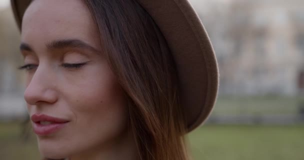 Hermosa joven con sombrero de pie al aire libre y mirando a un lado. Encantadora modelo femenina ajustando su cabello castaño. Fondo desenfocado. — Vídeo de stock