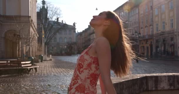 Vista lateral de la agradable mujer disfrutando del sol de la mañana con los ojos cerrados mientras está de pie en la calle de la ciudad. Adorable dama usando vestido de verano de moda. — Vídeos de Stock