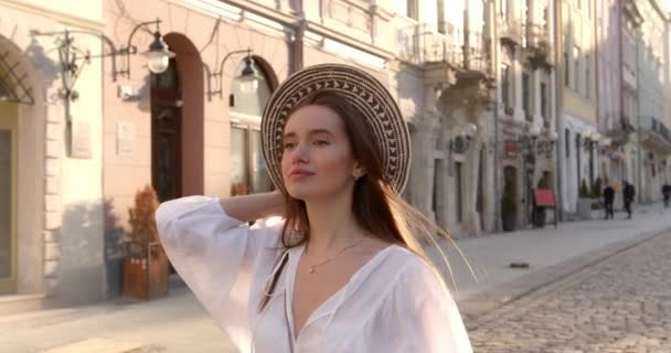 Modelo femenino feliz en elegante sombrero de verano y vestido blanco caminando tranquilamente en la calle de la ciudad. Encantadora mujer con cabello castaño disfrutando de días cálidos al aire libre. — Vídeos de Stock