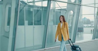 Sarı ceketli, kot pantolonlu, çantalı ve akıllı telefonlu güzel bir bayan modern havaalanı yakınlarında yürüyor. İş gezisi kavramı. 
