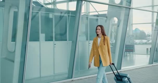 黄色のジャケットとブルーのジーンズの愛らしい若い女性は 現代の空港近くのスーツケースやスマートフォンで歩いています 出張の概念 — ストック動画