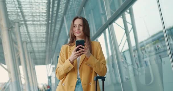 拿着智能手机站在现代机场附近靠在手提箱上微笑的女企业家 工作旅行的女商务人士 — 图库视频影像