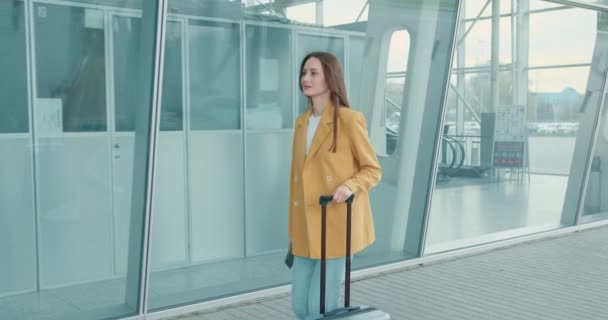 带着灰色手提箱的成功的女商人走在机场附近 手里拿着现代智能手机 穿着时髦的黄色夹克和蓝色牛仔裤的年轻女士 — 图库视频影像