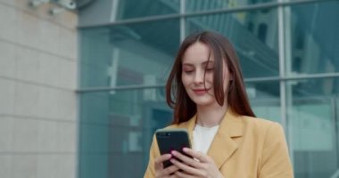 Kahverengi saçlı hoş bir bayan şehir binasının yakınında yürüyor ve modern akıllı telefon kullanıyor. Sarı ceket giyen sevimli bir kadın.. 