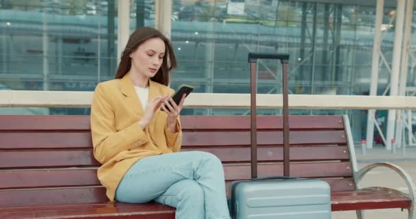 迷人的年轻女士穿着黄色夹克 坐在户外木制长椅上 使用现代智能手机 一个漂亮的女人 灰色的手提箱在机场附近等商务航班 — 图库视频影像