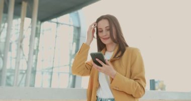 Sarı ceketli ve kot pantolonlu güzel bayan şehir binasının yanında duruyor ve modern akıllı telefon kullanıyor. Ofis dışındaki e-postaları kontrol eden hoş bir kadın.. 