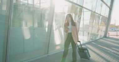 Resmi kıyafetler ve gözlüklü meşgul genç bir kadın havaalanı yakınlarında bavul ve akıllı telefonla yürüyor. İş kadını yurtdışında çalışıyor.. 