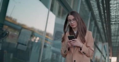 Güzel bayan girişimci şehir caddesinde yürürken kişisel cep telefonuna mesaj okuyor. Gözlüklü genç iş kadını, ofis dışında çalışmak için modern akıllı telefon kullanıyor..