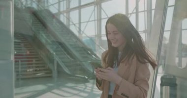Kafkasyalı genç iş kadını modern cam binanın yanında yürüyor ve akıllı telefondan internette sörf yapıyor. Kahverengi saçlı gülümseyen bayan şık ofis kıyafetleri giyiyor.. 