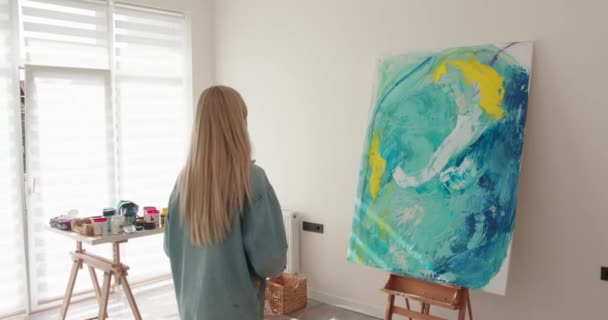 長いブロンドの髪を持つ才能のある女性画家のバックビューは、アートスタジオに立って抽象的な図面を見て。若い女性推定彼女の傑作といくつかの色を追加. — ストック動画
