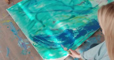 Sarı saçlı, beyaz tuvalde spatulayla renkleri karıştıran genç bir kadın. Sıradan giysiler içinde kadın ressam yerde oturuyor ve çizim yapıyor.. 