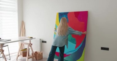 Sarı saçlı güzel genç bir kadın iş yerinde soyut boyalı büyük bir tuval taşıyor. Modern stüdyoda rengarenk paleti olan gündelik kıyafet çizen mutlu kadın sanatçı..