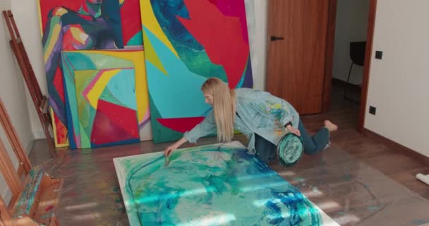 Inspirado jovem senhora em roupas casuais pintura em tela grande com escova e paleta de cores. Artista feminina sentada no chão no estúdio de arte e desfrutando de processo criativo. — Vídeo de Stock