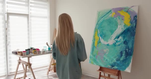 Back view of young woman creating masterpiece on canvas at bright studio. Colorful abstract pattern on easel. Fantasy of talented artist. — Wideo stockowe