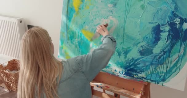 Vista posteriore della donna vestita con disinvoltura seduta davanti al cavalletto e disegno astratto pittura. Artista donna ispirata utilizzando spatola e tavolozza dei colori al lavoro. — Video Stock