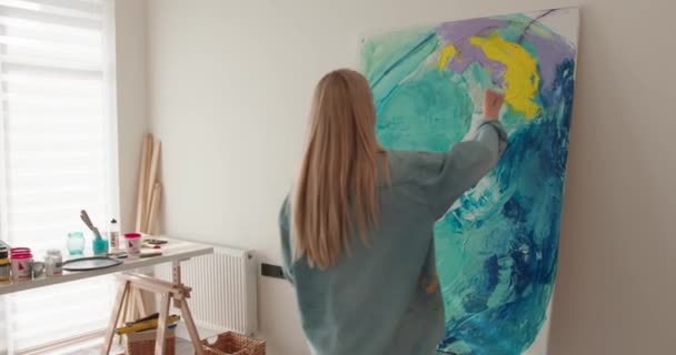 Rückenansicht einer jungen Frau in lässigem Outfit, die mit den Fingern auf der Staffelei zeichnet. Inspirierte Künstlerin mischt Farben auf Leinwand im Kunstatelier. — Stockvideo