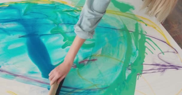 Joven pintora usando paleta de colores y pincel mientras dibuja un patrón abstracto sobre lienzo. Primer plano de la mujer con el pelo rubio sentado en el suelo en el estudio de arte. — Vídeos de Stock