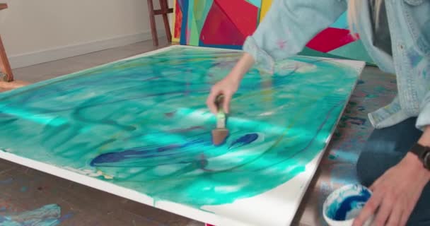 Geïnspireerde jonge vrouw die op de vloer zit en spatel en blauwe verf gebruikt terwijl ze abstracte tekeningen maakt op doek. Sluiten van het werkproces in het atelier. — Stockvideo