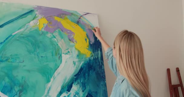 Caucasian young woman in casual wear creating drawing on easel at bright studio. Talented artist using brush and color palette for painting. — Stock video