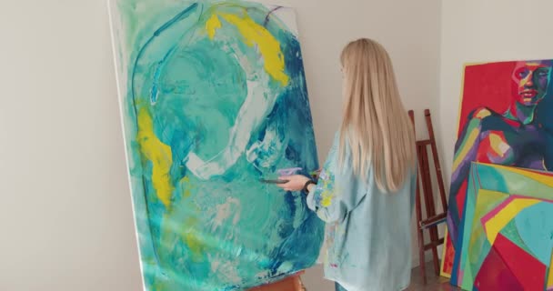 Glückliche junge Frau in Freizeitkleidung mit Pinsel und Farbpalette, während sie im Atelier auf Staffelei zeichnet. Künstlerin mit blonden Haaren genießt kreatives Hobby in der Freizeit. — Stockvideo