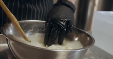 Siyah eldivenli bir adam peyniri karıştırmak için ahşap spatula kullanıyor.