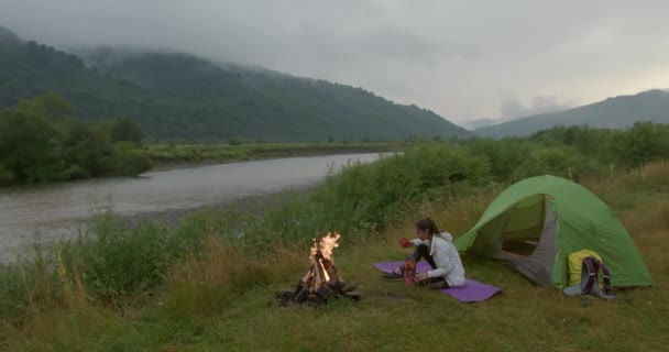 Kvinna sitter nära tält på kullen och njuter av utsikten över floden — Stockvideo