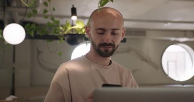 Kafe masasında dizüstü bilgisayarı ve cep telefonu olan gülümseyen adam.