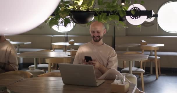 Mann hält Handy in der Hand, während er mit Laptop im Café sitzt — Stockvideo