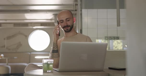 Positiv man skriva på bärbar dator och prata på mobil på café — Stockvideo