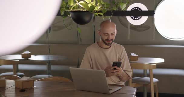 Hombre positivo usando el ordenador portátil y el teléfono celular en la mesa de café — Vídeos de Stock