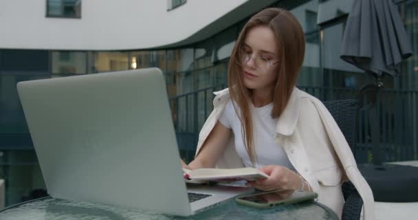 Mujer joven sentada al aire libre con portátil y libro de lectura — Vídeos de Stock