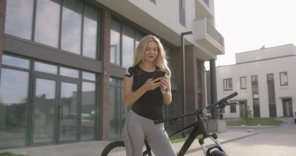 Kobieta w odzieży sportowej siedzi na rowerze i używa telefonu komórkowego — Wideo stockowe