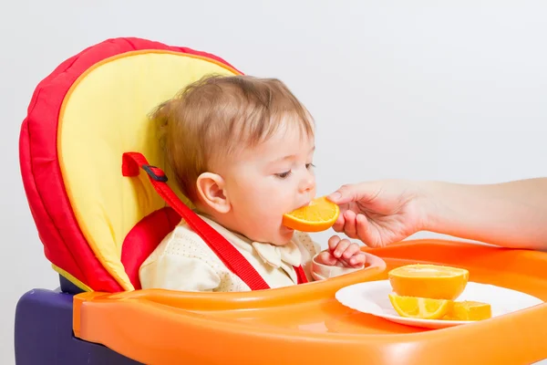 Baby zit in kinderstoel met sinaasappel. — Stockfoto