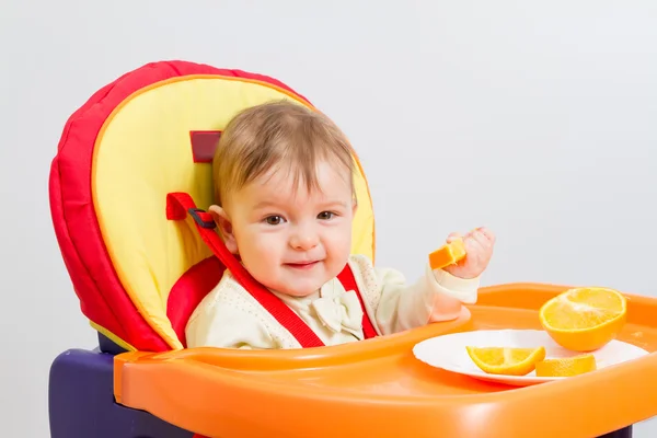 Baby zit in kinderstoel met sinaasappel. — Stockfoto